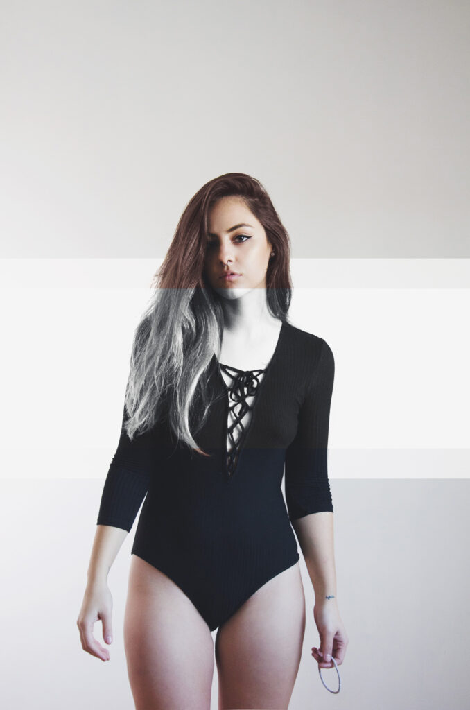 Laura Adriana photo by Ivan Beoulve. A model with long wavy hair stands against a plain background wearing a black bodysuit with a crisscross lace-up front.